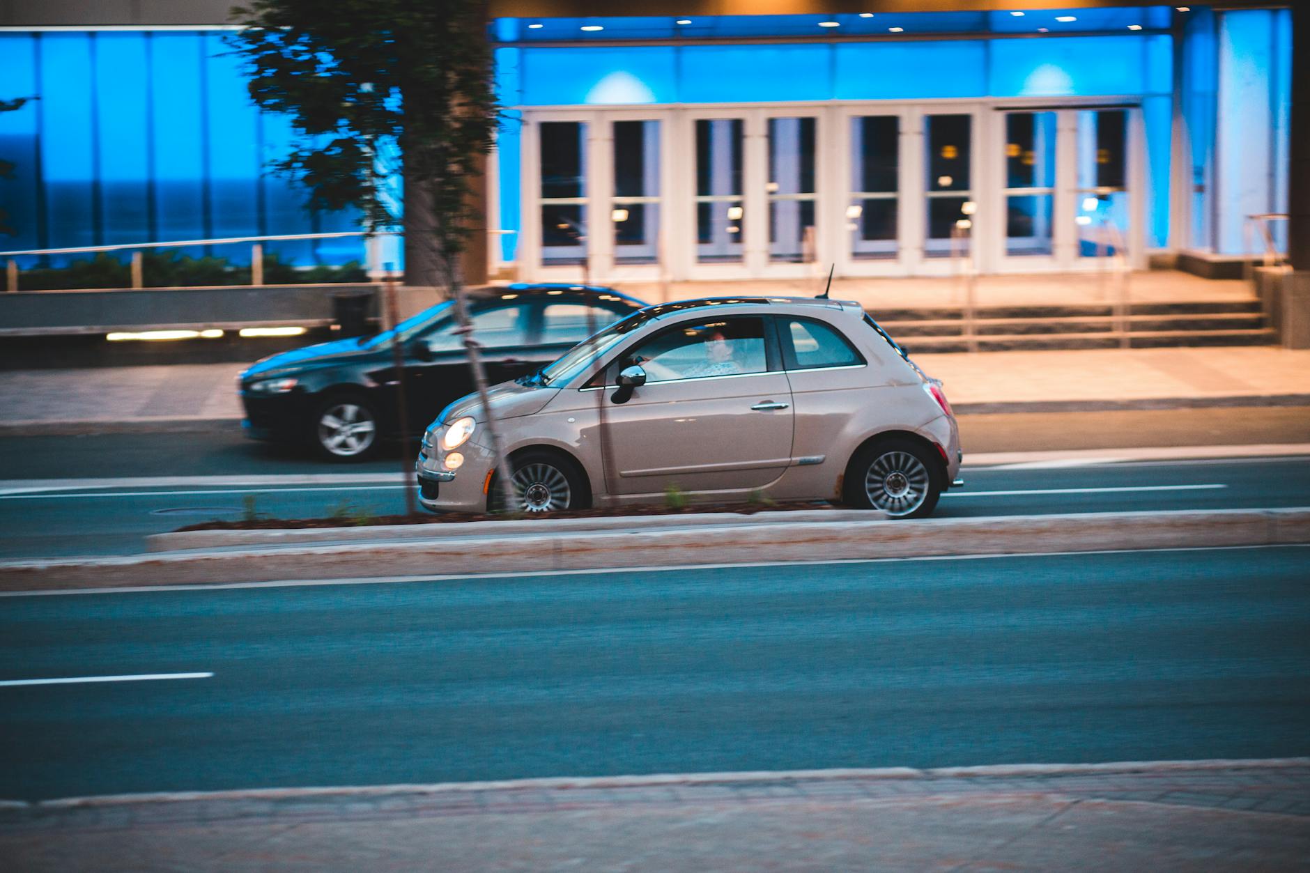 Białe Fiat 500 jadące nocą obok nowoczesnego, niebiesko podświetlonego biurowca